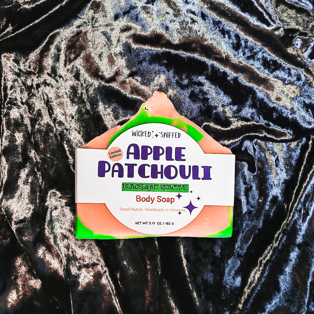 A bar of Apple Patchouli Body Soap by Wicked Sniffer, featuring a white label with purple text and green accents, resting on a dark, crinkled velvet fabric background.