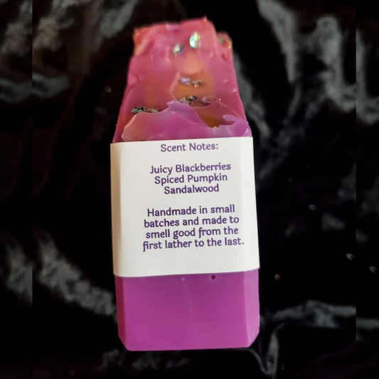 A handmade rectangular bar of soap in pink and purple with glittery details, wrapped in a white paper label that reads 'Scent Notes: Juicy Blackberries Spiced Pumpkin Sandalwood' and 'Handmade in small batches and made to smell good from the first lather to the last.'.