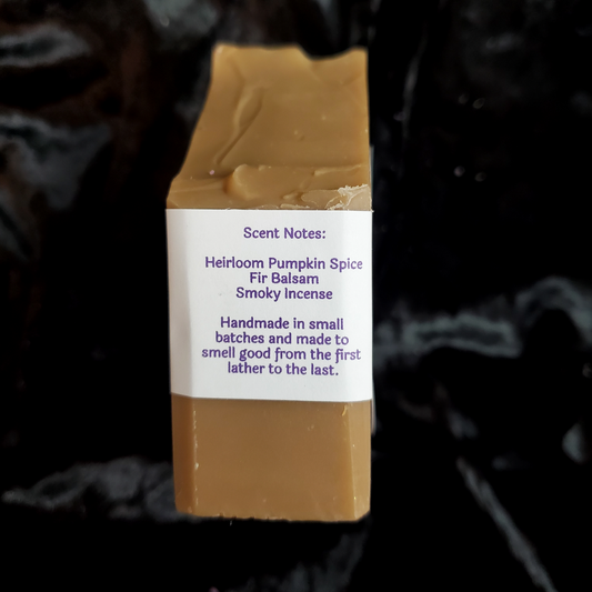 A rectangular bar of soap with a white paper label reading scent notes for 'Heirloom Pumpkin Spice Fir Balsam Smoky Incense,' photographed against a dark black background.