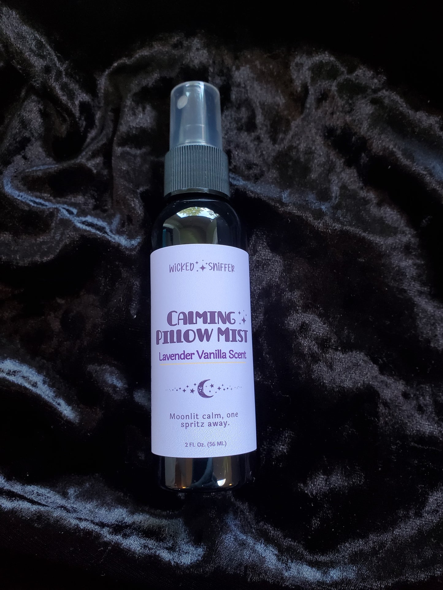 A 2 fl oz bottle Calming Pillow Mist in Lavender Vanilla Scent, featuring a purple label with purple text, set against a dark, textured velvet background.
