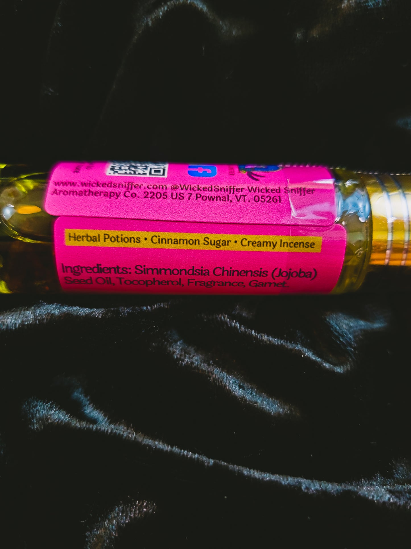 A close-up of a Wicked Sniffer brand herbal potion bottle with a bright pink label, resting on a black textured fabric background.