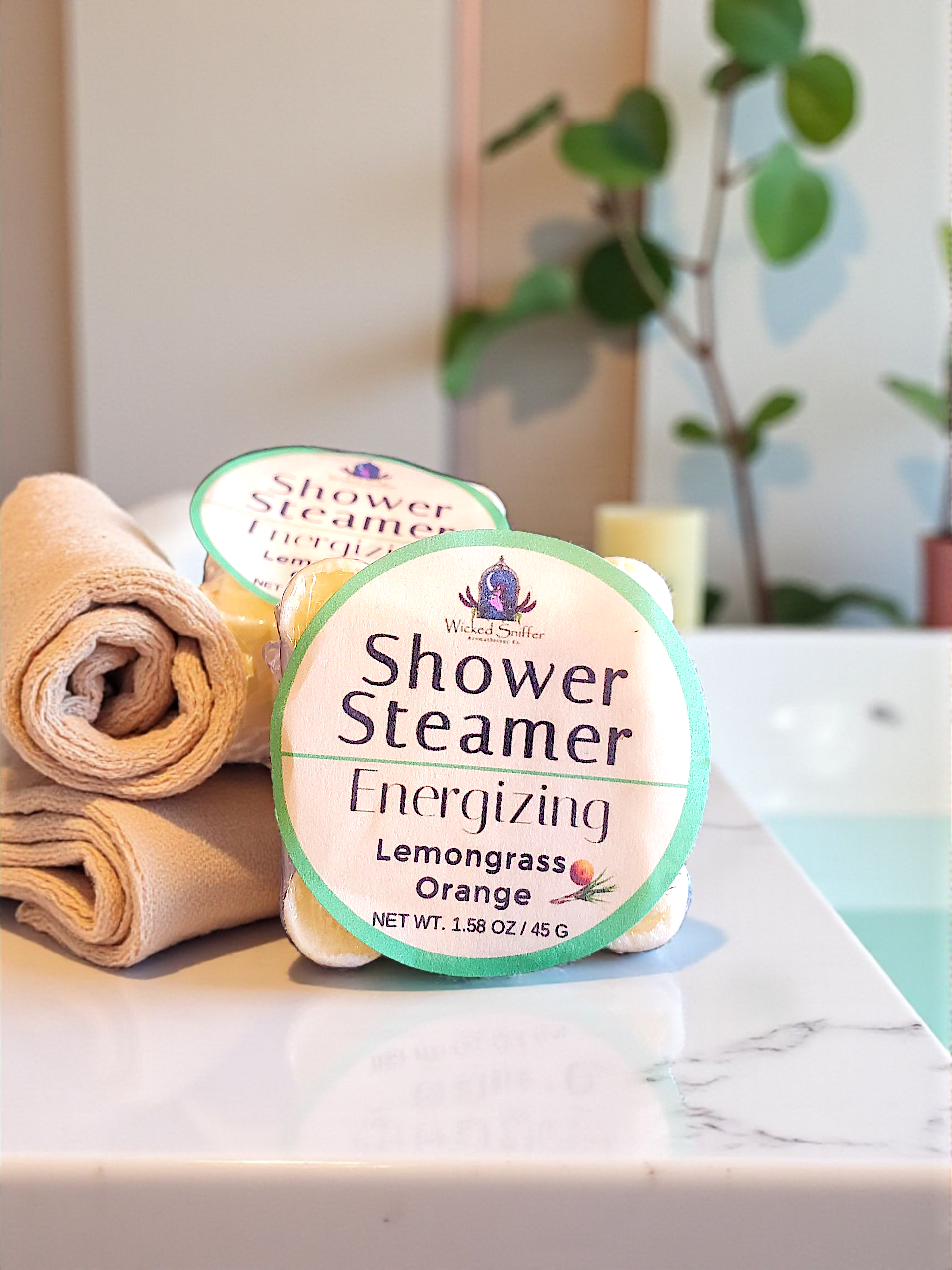Two Shower Steamers on a marble surface with a towel and plant in the background