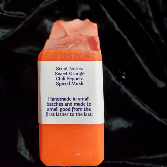 A rectangular bar of handmade soap in a vibrant orange color, featuring a white paper label with scent notes and handmade details. The soap is set against a dark, draped black fabric background.