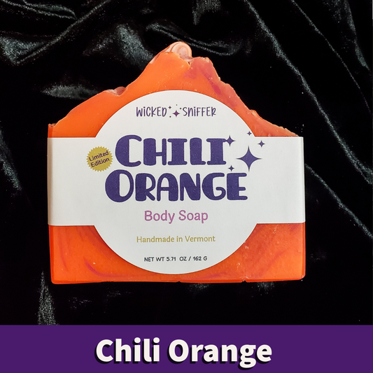 A handcrafted chili orange body soap bar with a white paper label reading 'Wicked Sniffer Chili Orange Body Soap' on a black velvet background, with a purple banner at the bottom labeled 'Chili Orange'.