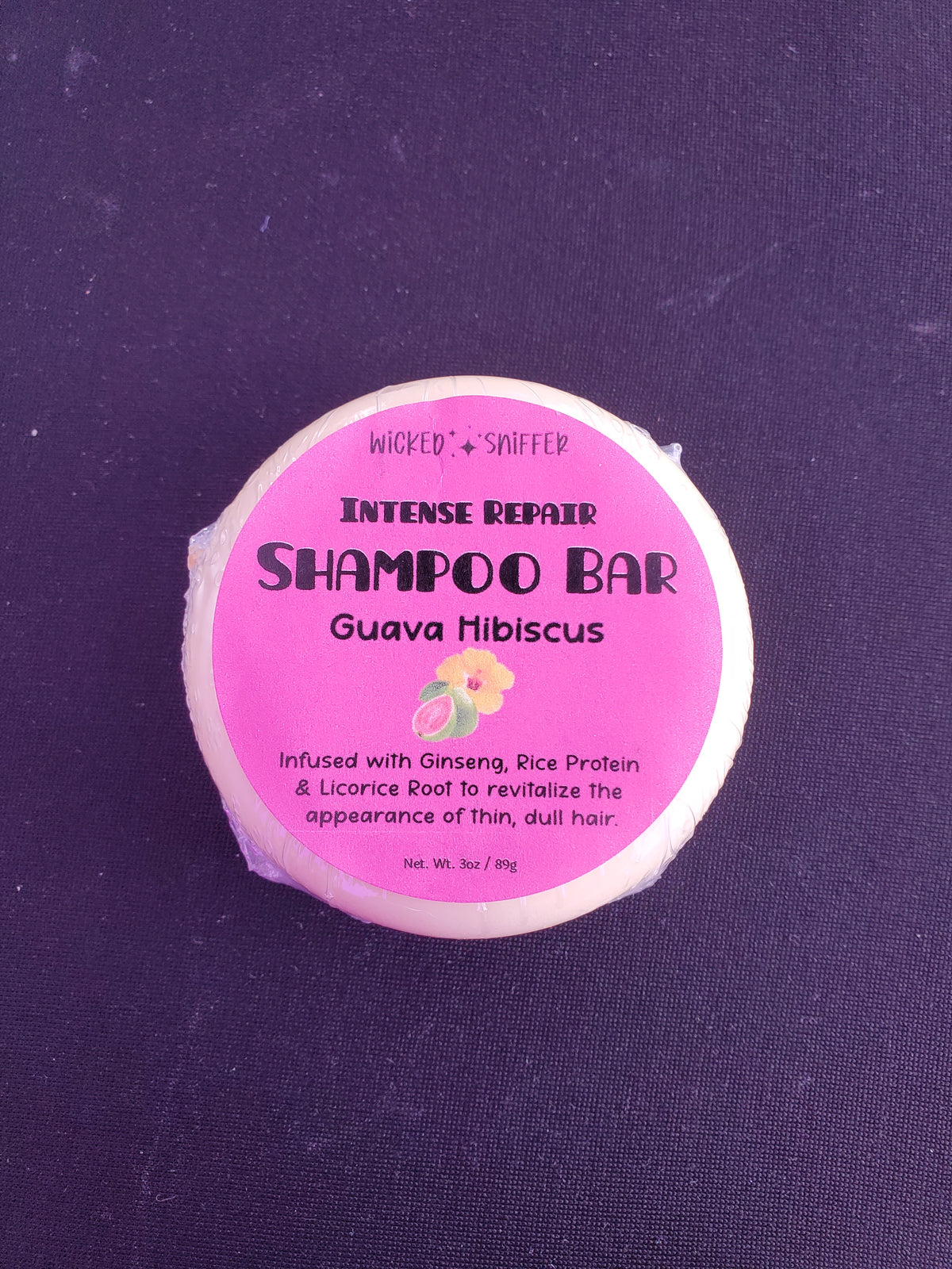 A round shampoo bar wrapped in clear plastic with a pink label reading 'Wicked Sniffer Intense Repair Shampoo Bar Guava Hibiscus' on a dark textured background.