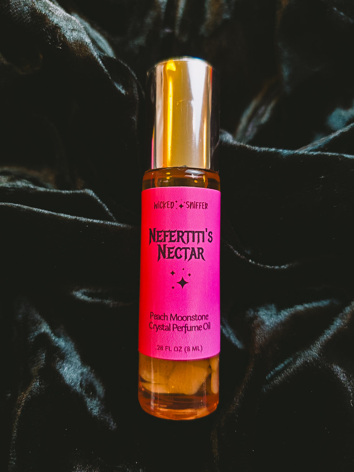 A 10 mL bottle of Wicked & Sniffed Nefertiti's Nectar Peach Moonstone Crystal Perfume Oil, featuring a bright pink label and a metallic gold cap, set against a draped black velvet background.