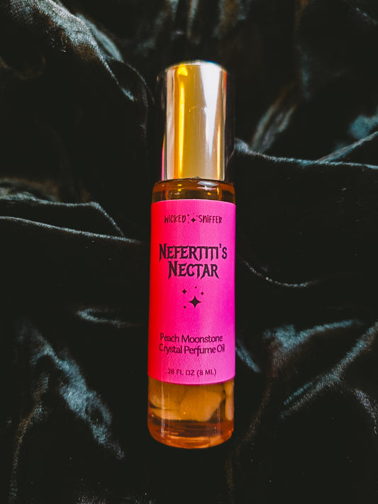A 10 mL bottle of Wicked & Sniffed Nefertiti's Nectar Peach Moonstone Crystal Perfume Oil, featuring a bright pink label and a metallic gold cap, set against a draped black velvet background.