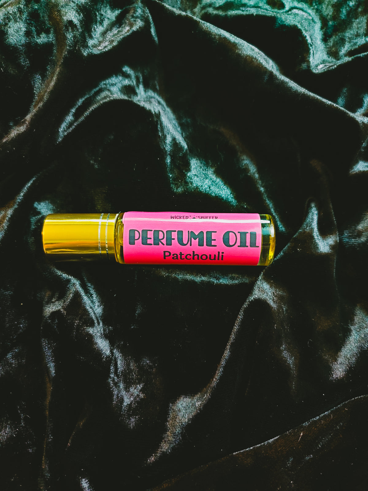 A perfume oil rollerball bottle with a bright pink label reading 'PERFUME OIL Patchouli' and a metallic gold cap, placed on a dark green velvet fabric background.