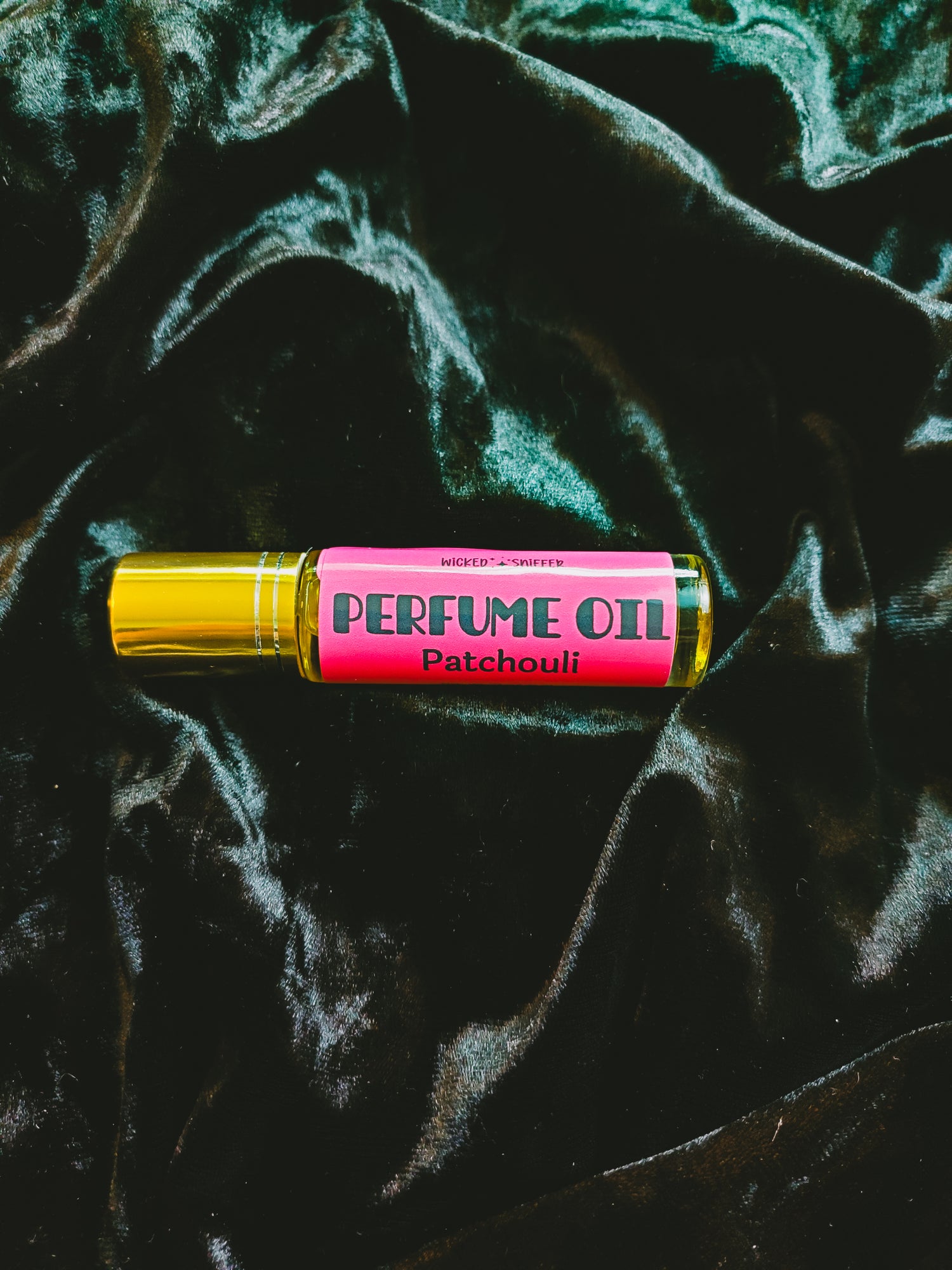 A perfume oil rollerball bottle with a bright pink label reading 'PERFUME OIL Patchouli' and a metallic gold cap, placed on a dark green velvet fabric background.