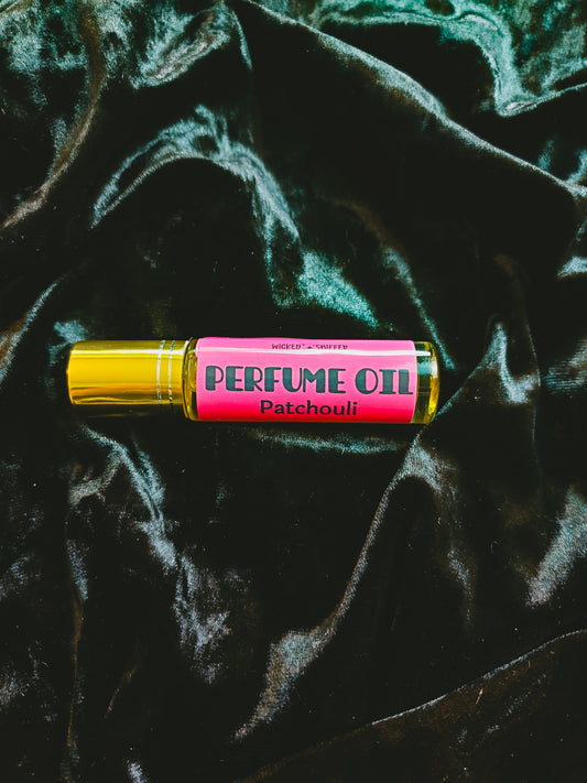 A perfume oil rollerball bottle with a bright pink label reading 'PERFUME OIL Patchouli' and a metallic gold cap, placed on a dark green velvet fabric background.
