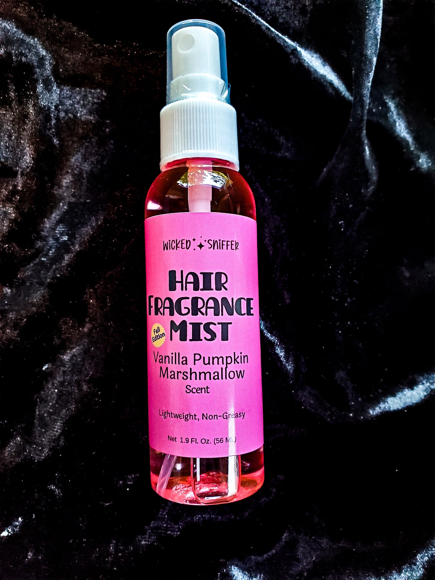 A pink bottle with a white cap and pink label that has black lettering that says Hair fragrance mist, on a black velvet background.