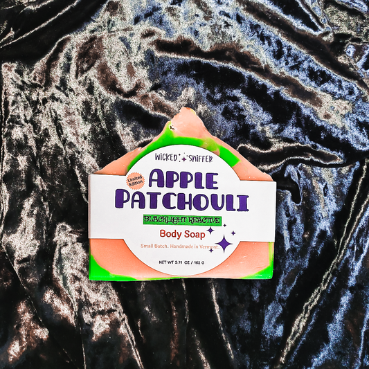 A bar of Apple Patchouli Body Soap by Wicked Sniffer, featuring a white label with purple text and green accents, resting on a dark, crinkled velvet fabric background.