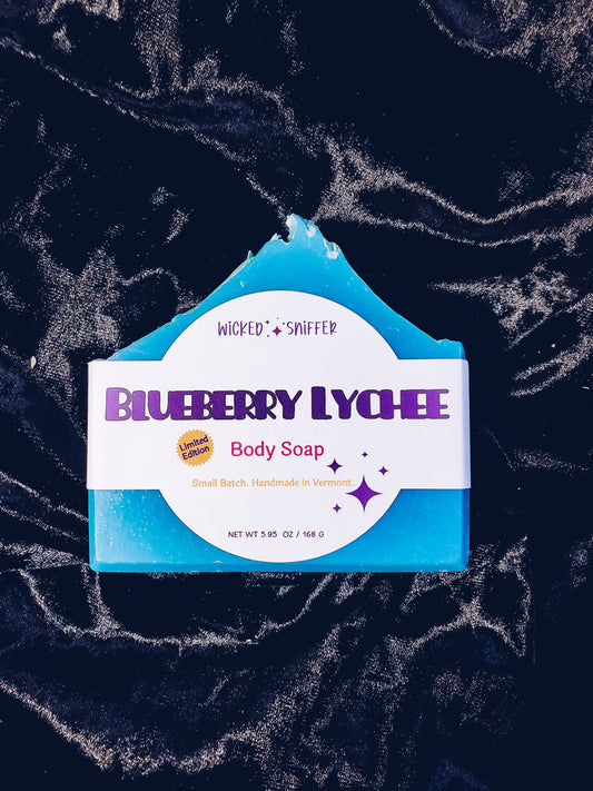 A bar of blueberry lychee body soap by Wicked & Sniffer, featuring a white label with purple text and a bright blue soap bar visible at the top and sides, set against a dark marbled background.