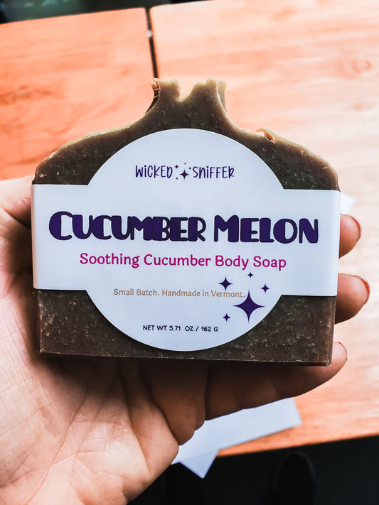 A hand holding a bar of Wicked Sniffer Cucumber Melon Soothing Cucumber Body Soap. The soap bar is green with visible speckles, wrapped in a white paper label with purple and pink text, set against a blurred wooden background.
