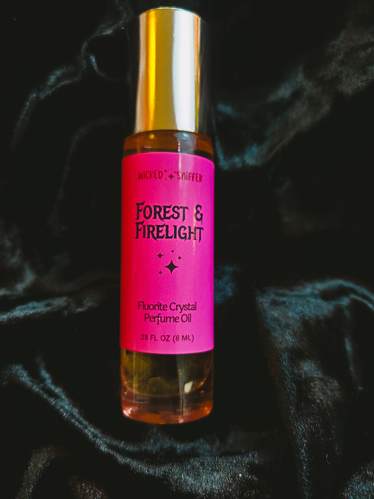 A roll-on bottle of Forest & Firelight Perfume Oil, featuring a bright pink label and a metallic gold cap, set against a dark, draped velvet fabric background.