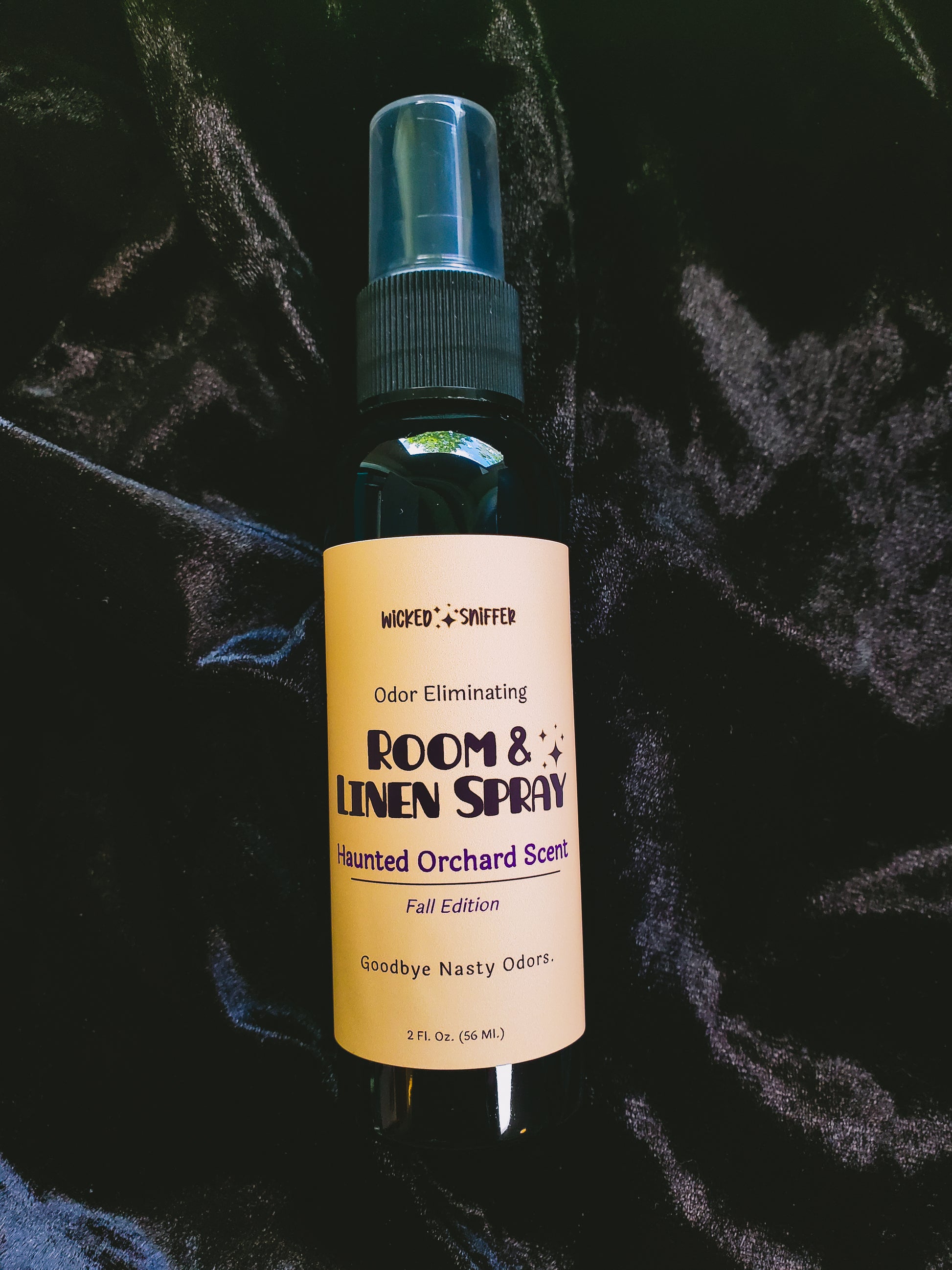 A 2 fl oz spray bottle of Room & Linen Spray in the 'Haunted Orchard Scent' fall edition, featuring a pale yellow label and a black spray nozzle, set against a dark, textured velvet background.
