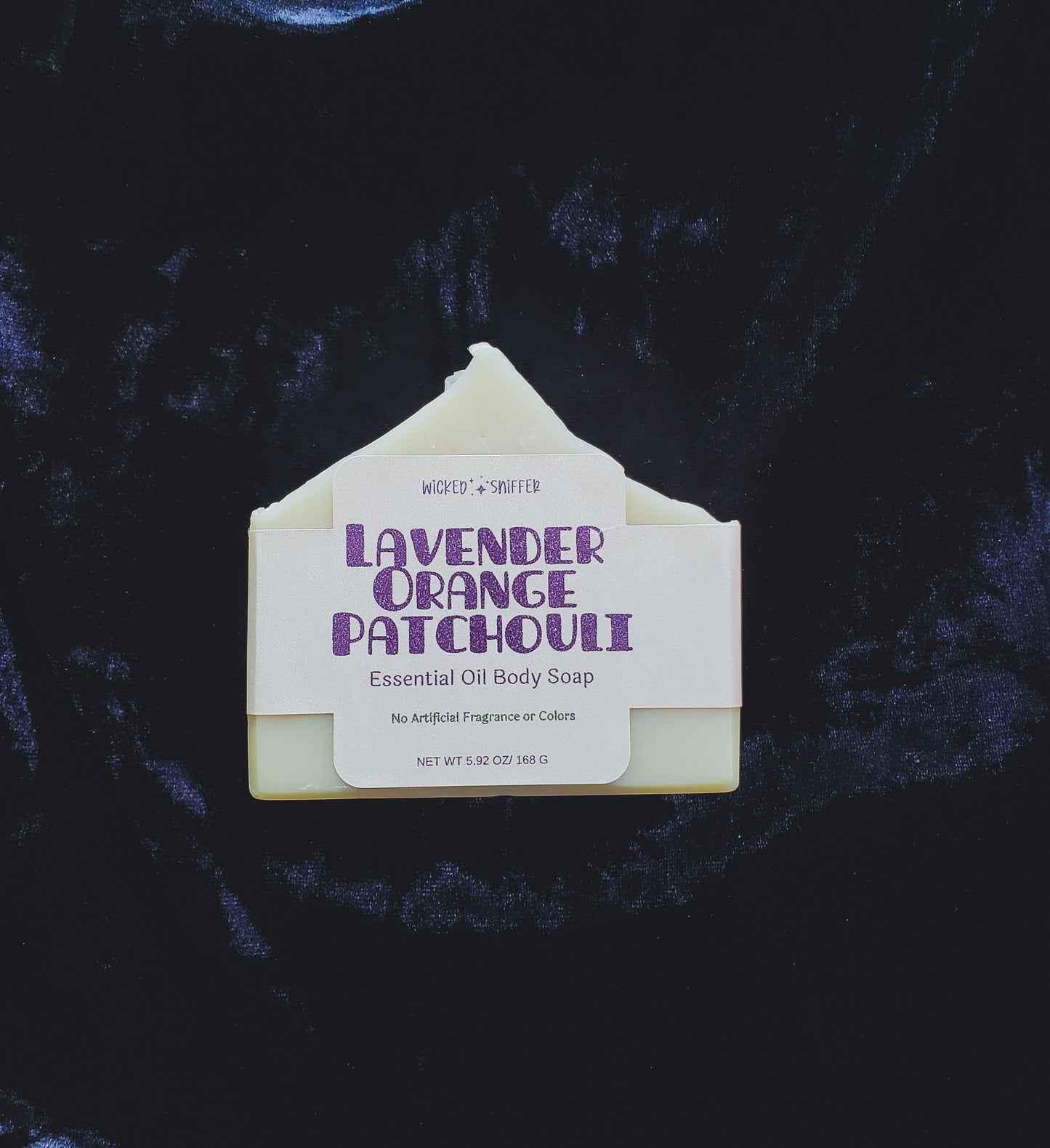 A single bar of Lavender Orange Patchouli Essential Oil Body Soap, shown with its paper label attached, set against a dark textured background.