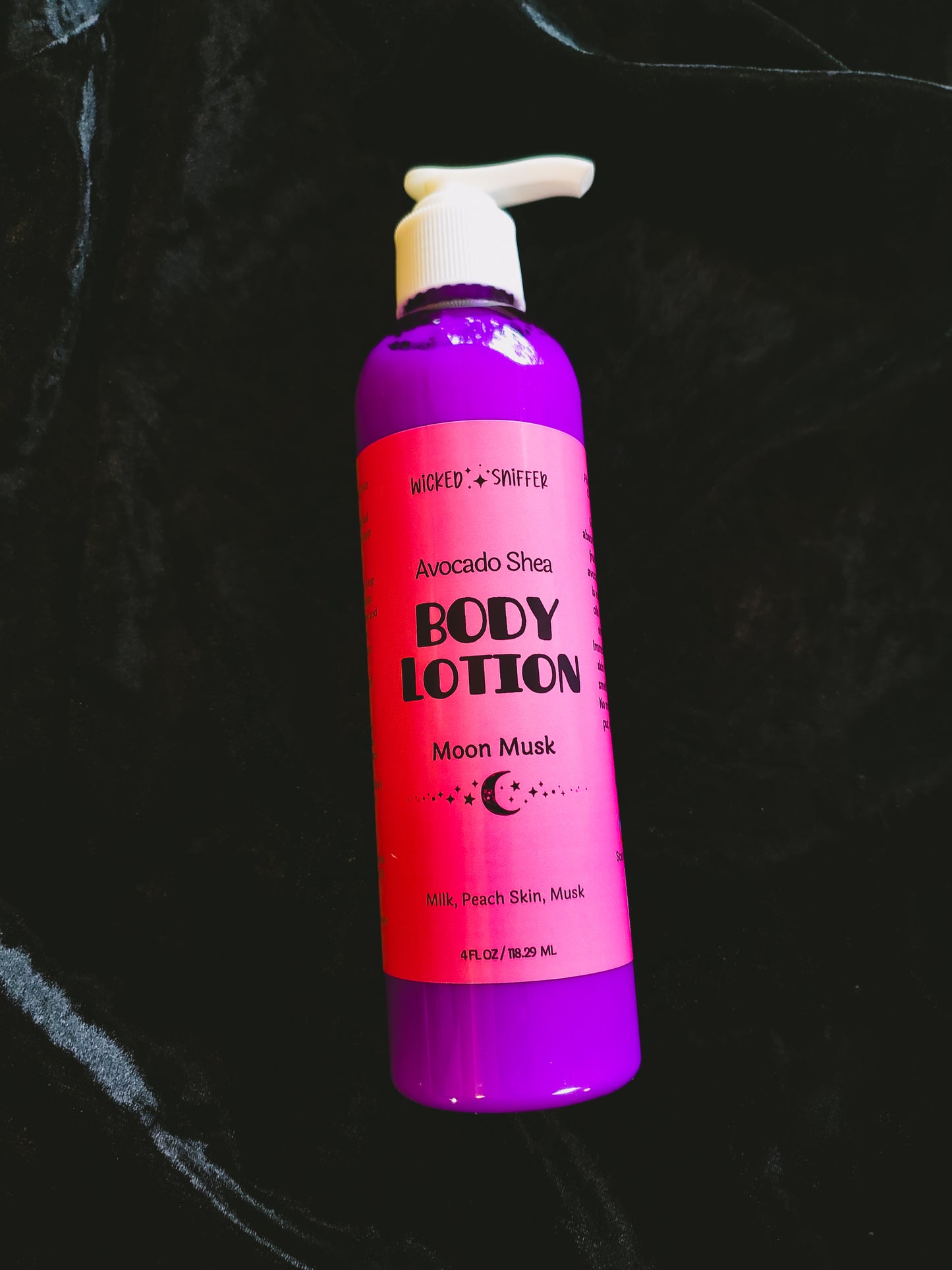 A bottle of Wicked Sniffer Avocado Shea Body Lotion in 'Moon Musk' scent, featuring a bright pink label and a purple bottle with a white pump dispenser, set against a dark textured background.