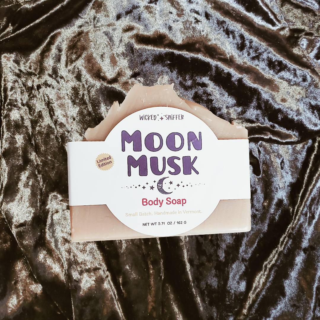 A bar of handmade body soap labeled 'Moon Musk' by Wicked Sniffer, placed on a shiny dark velvet fabric background.