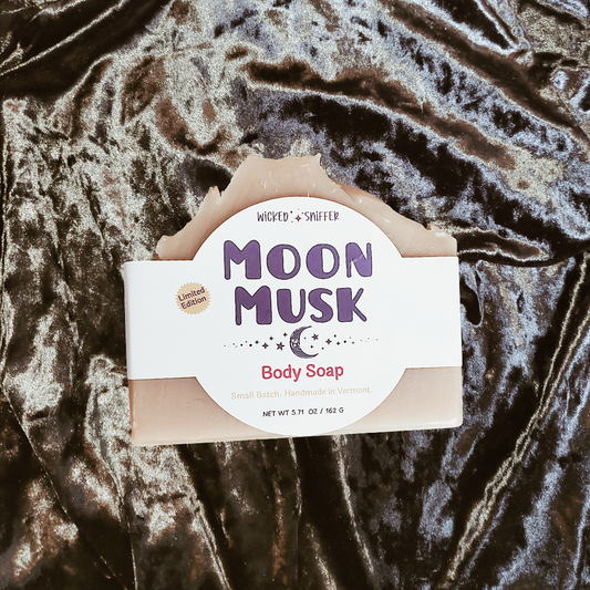 A bar of handmade body soap labeled 'Moon Musk' by Wicked Sniffer, placed on a shiny dark velvet fabric background.