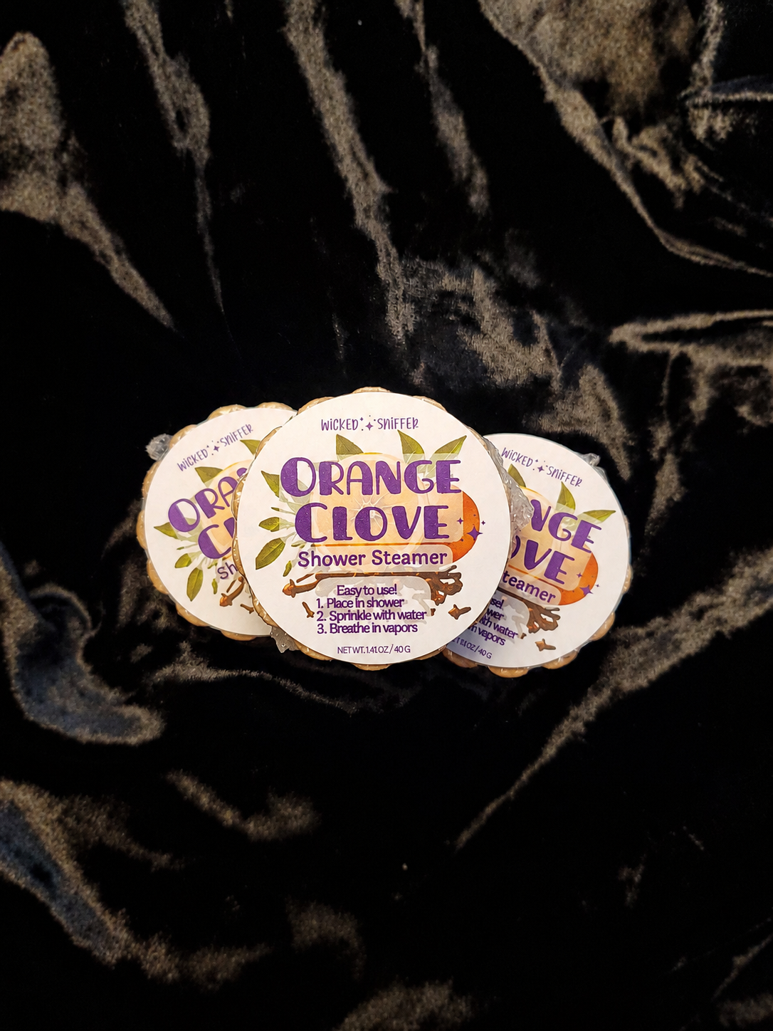 Orange Clove shower steamer packaging on a dark textured fabric background