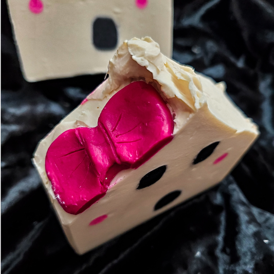 A close-up of a handmade soap bar shaped like a character face, featuring a beige base with a shiny hot pink bow and black facial features, set against a black textured background.