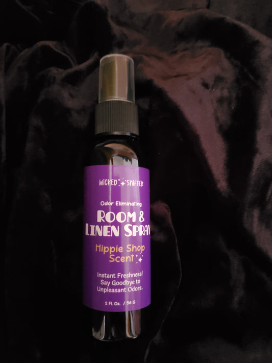 A small room spray bottle with a purple label reading 'ROOM & LINEN SPRAY' and 'Hippie Shop Scent' on a dark, textured fabric background.