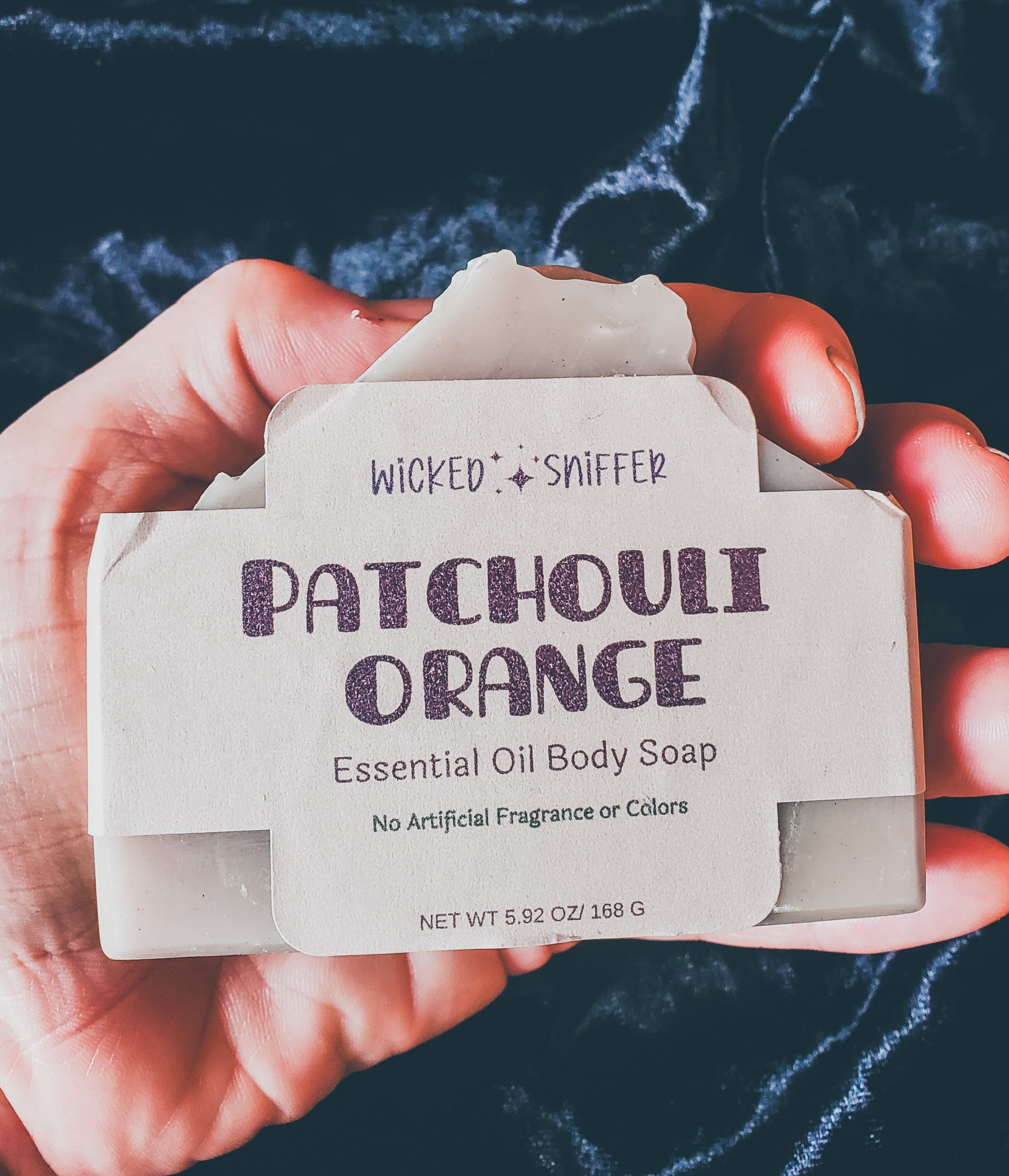Hand holding a tan bar of patchouli orange soap on a black background