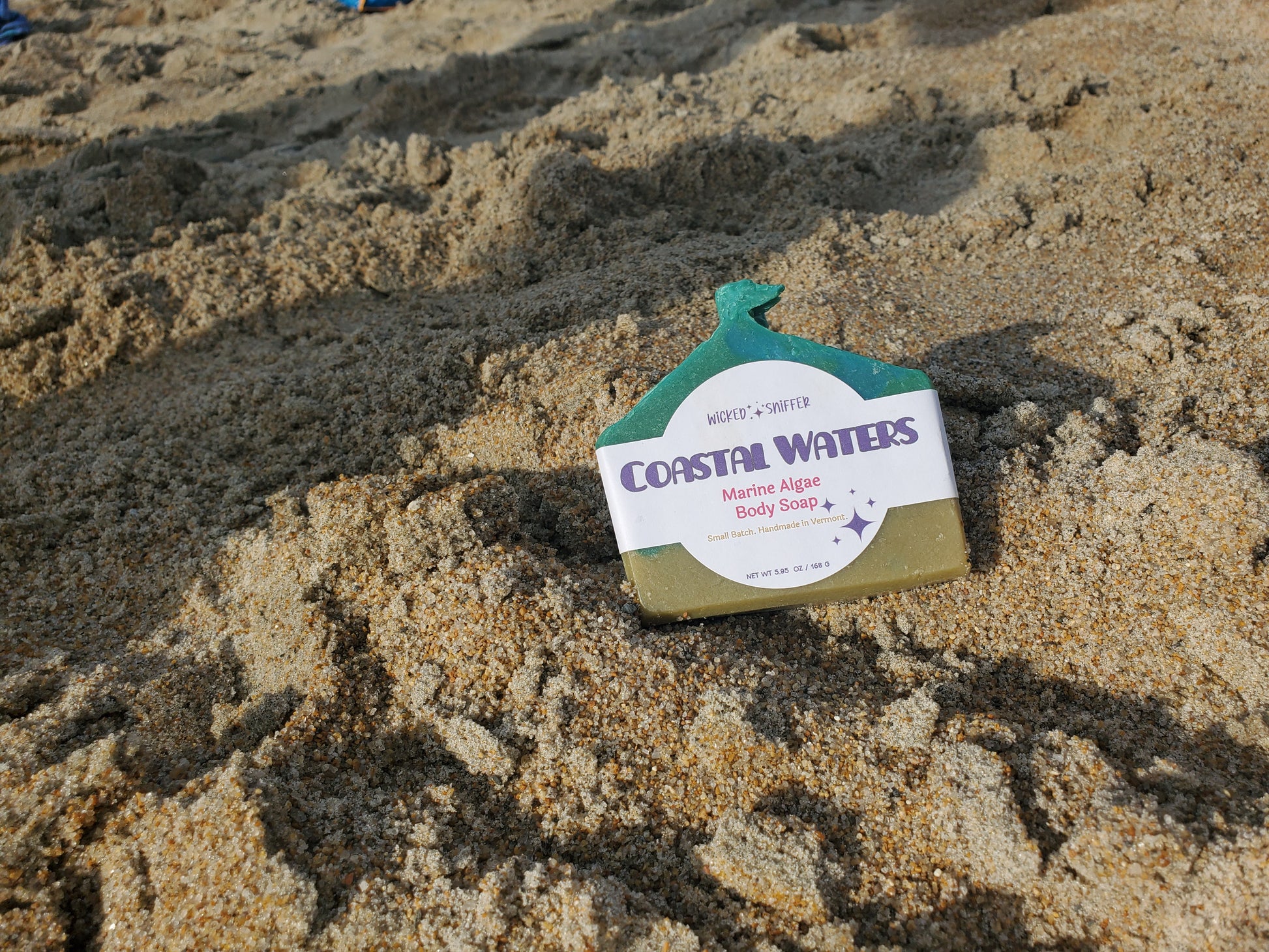 A bar of Coastal Waters Marine Algae Body Butter sits on sandy beach ground, with the product label clearly visible against the tan sand.