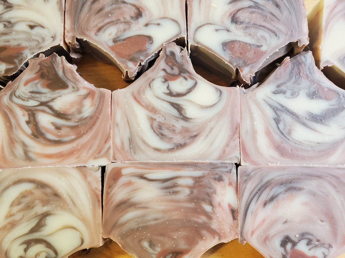Close-up of sliced artisanal soap bars with a marbled swirl pattern of dusty pink, cream, and gray on a wooden surface.