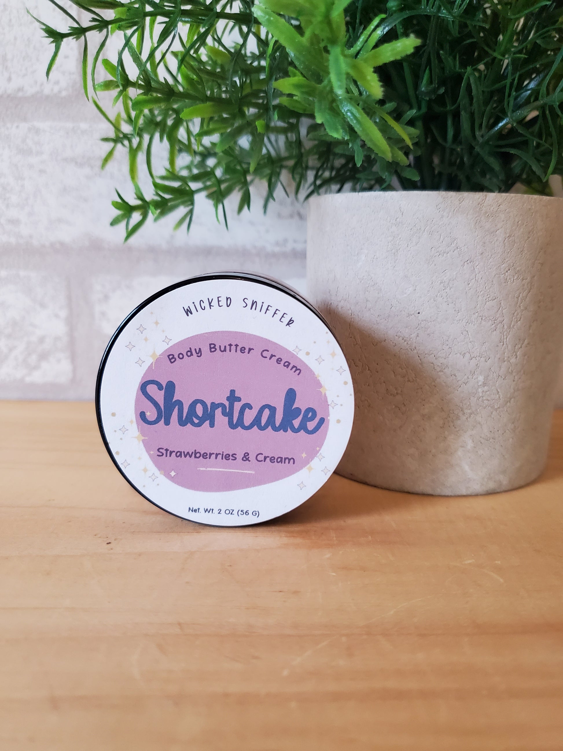 Body butter container labeled 'Shortcake' on a wooden surface with greenery.