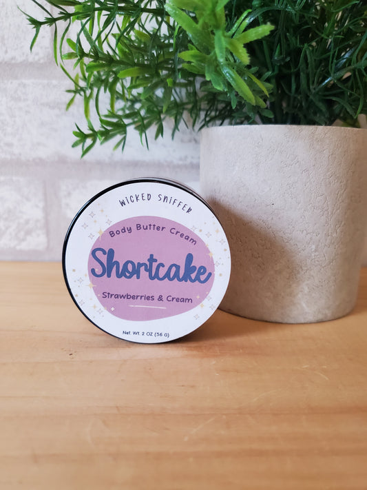 Body butter container labeled 'Shortcake' on a wooden surface with greenery.