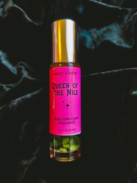 A 2.6 fl oz rollerball bottle of Wicked & Sniffed 'Queen of the Nile' Ruby Zoisite Crystal Perfume Oil, featuring a bright pink label and a metallic gold cap, set against a dark, draped velvet background.