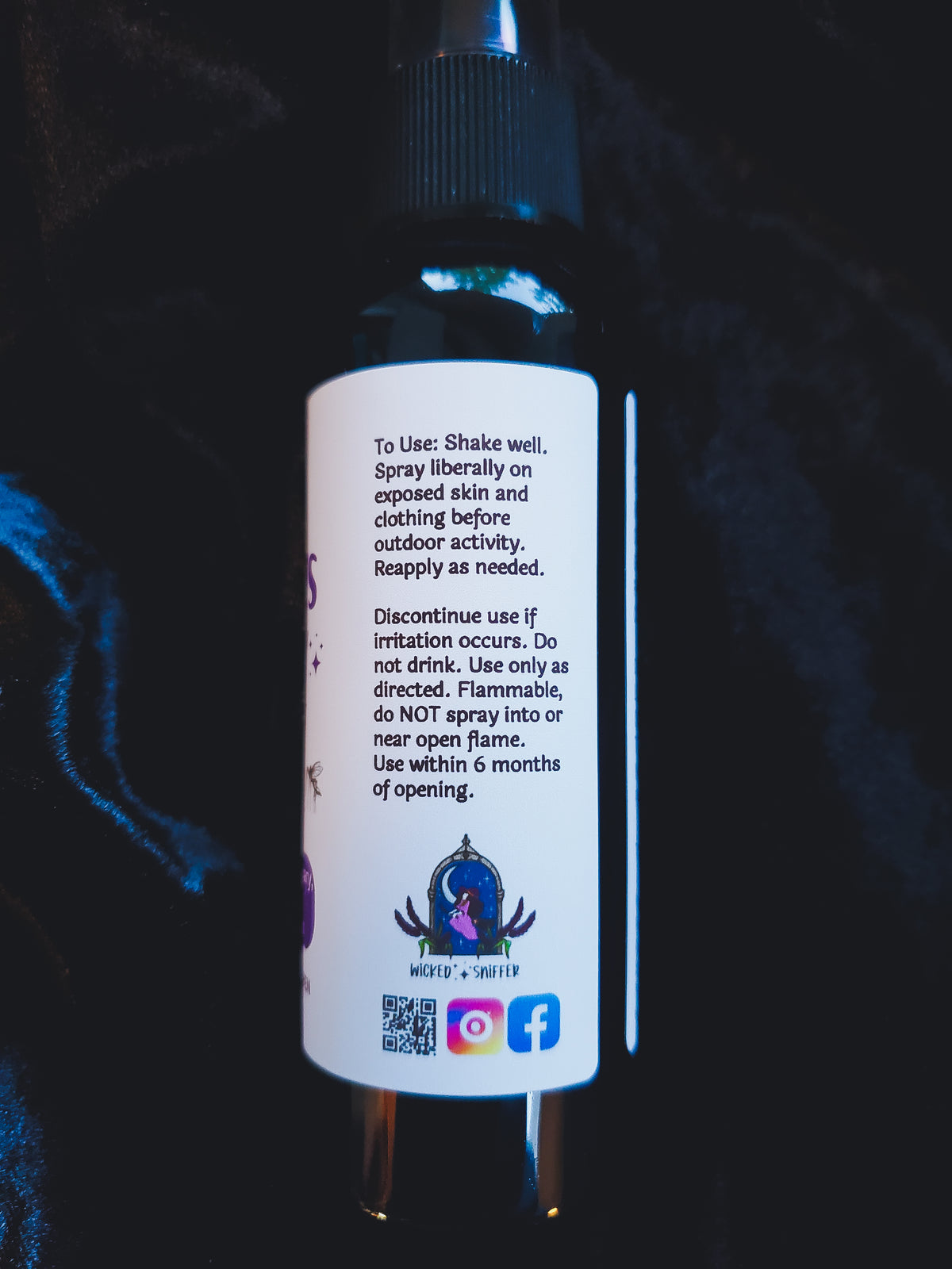 A close-up of the back of a black spray bottle with a white label, showing usage instructions and a small logo, set against a dark textured background.
