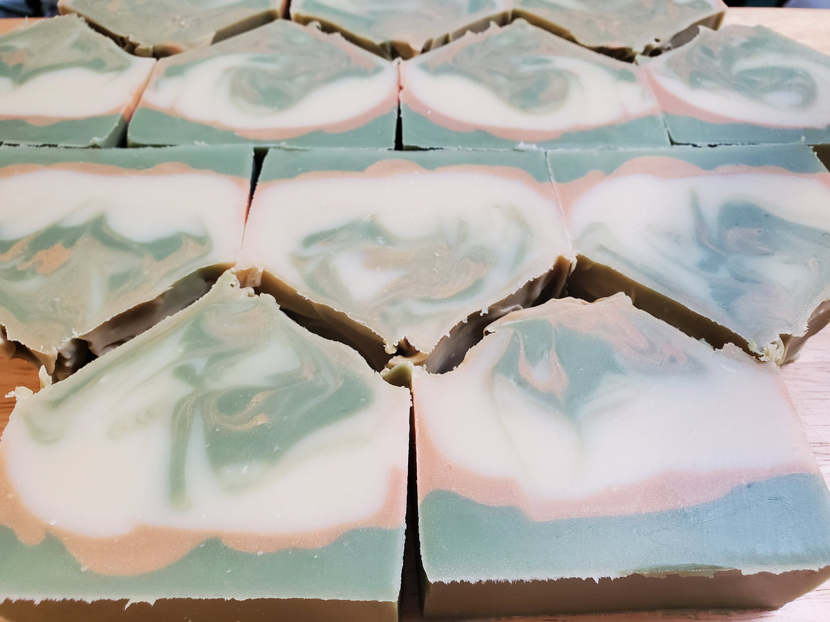 Multiple rectangular bars of handmade soap arranged in rows on a light wooden surface. Each bar has a swirled marbled pattern with white, sage green, dusty pink, and brown layers.