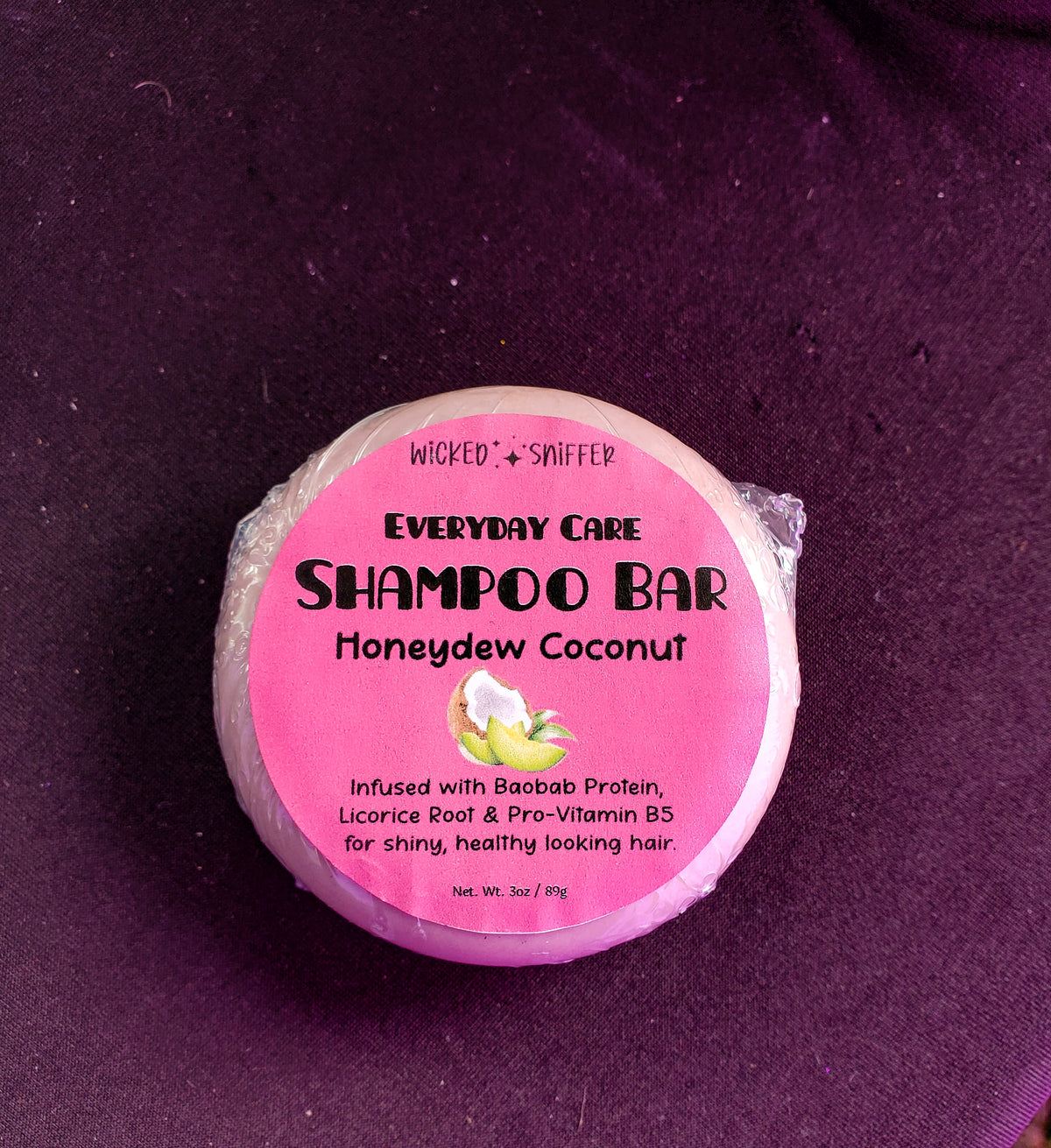 A round shampoo bar wrapped in clear plastic with a pink label reading 'Wicked Sniffer Everyday Care Shampoo Bar Honeydew Coconut,' placed on a dark purple fabric background.