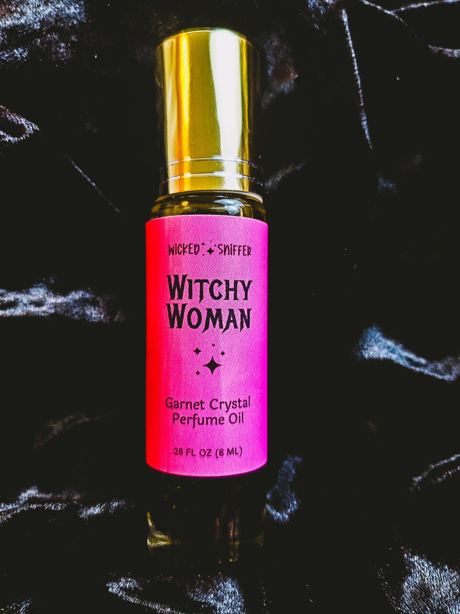 A 2 FL OZ bottle of Wicked by Sniffer Witchy Woman Garnet Crystal Perfume Oil, featuring a bright pink label and a shiny gold cap, set against a dark, textured background.