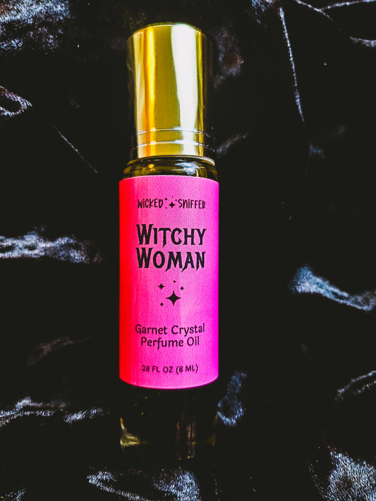 A 2 FL OZ bottle of Wicked by Sniffer Witchy Woman Garnet Crystal Perfume Oil, featuring a bright pink label and a shiny gold cap, set against a dark, textured background.