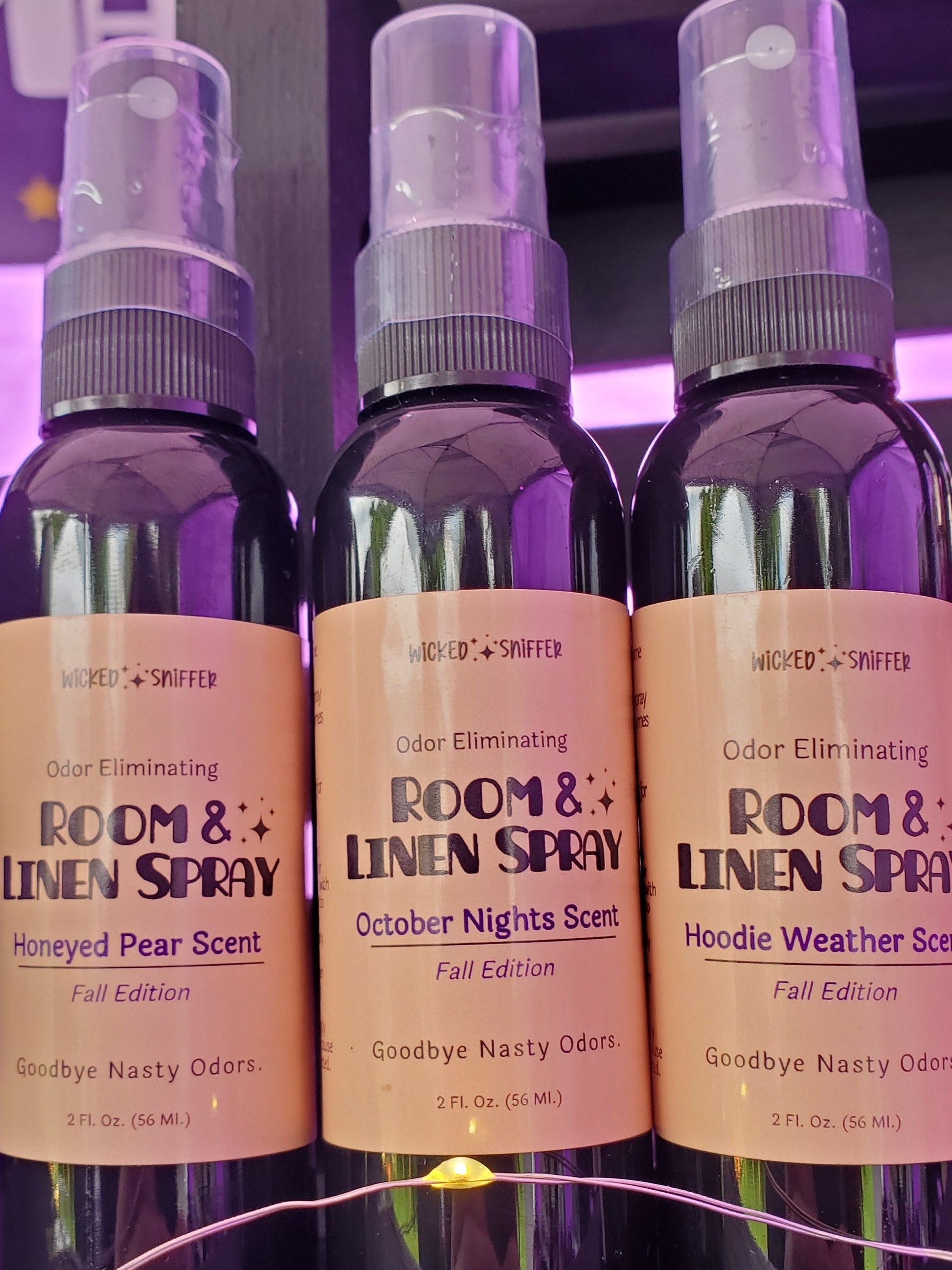 Three dark plastic room and linen spray bottles with tan labels are displayed on a dark wooden surface. The center bottle is labeled 'ROOM & LINEN SPRAY' in 'Honeyed Pear Scent', flanked by 'October Nights Scent' and 'Hoodle Weather Scent' bottles, with a purple-lit background.