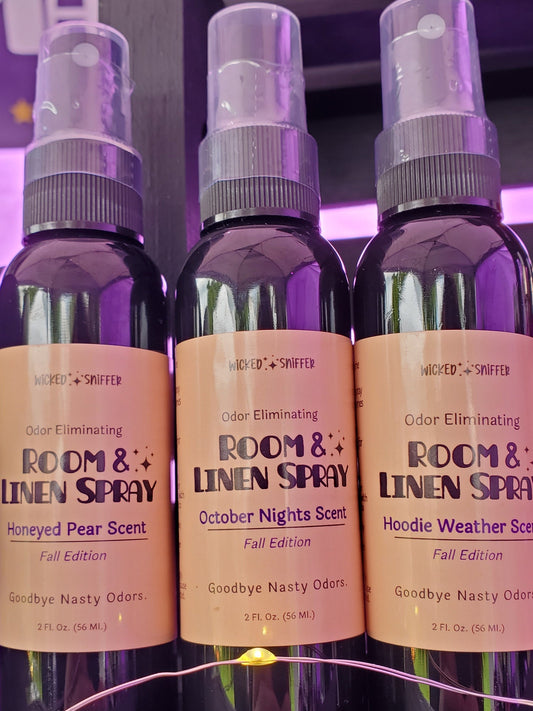 Three dark plastic room and linen spray bottles with tan labels are displayed on a dark wooden surface. The center bottle is labeled 'ROOM & LINEN SPRAY' in 'Honeyed Pear Scent', flanked by 'October Nights Scent' and 'Hoodle Weather Scent' bottles, with a purple-lit background.