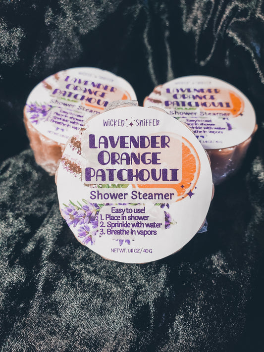 Three round shower steamers labeled 'Lavender Orange Patchouli' arranged on a dark textured fabric background.