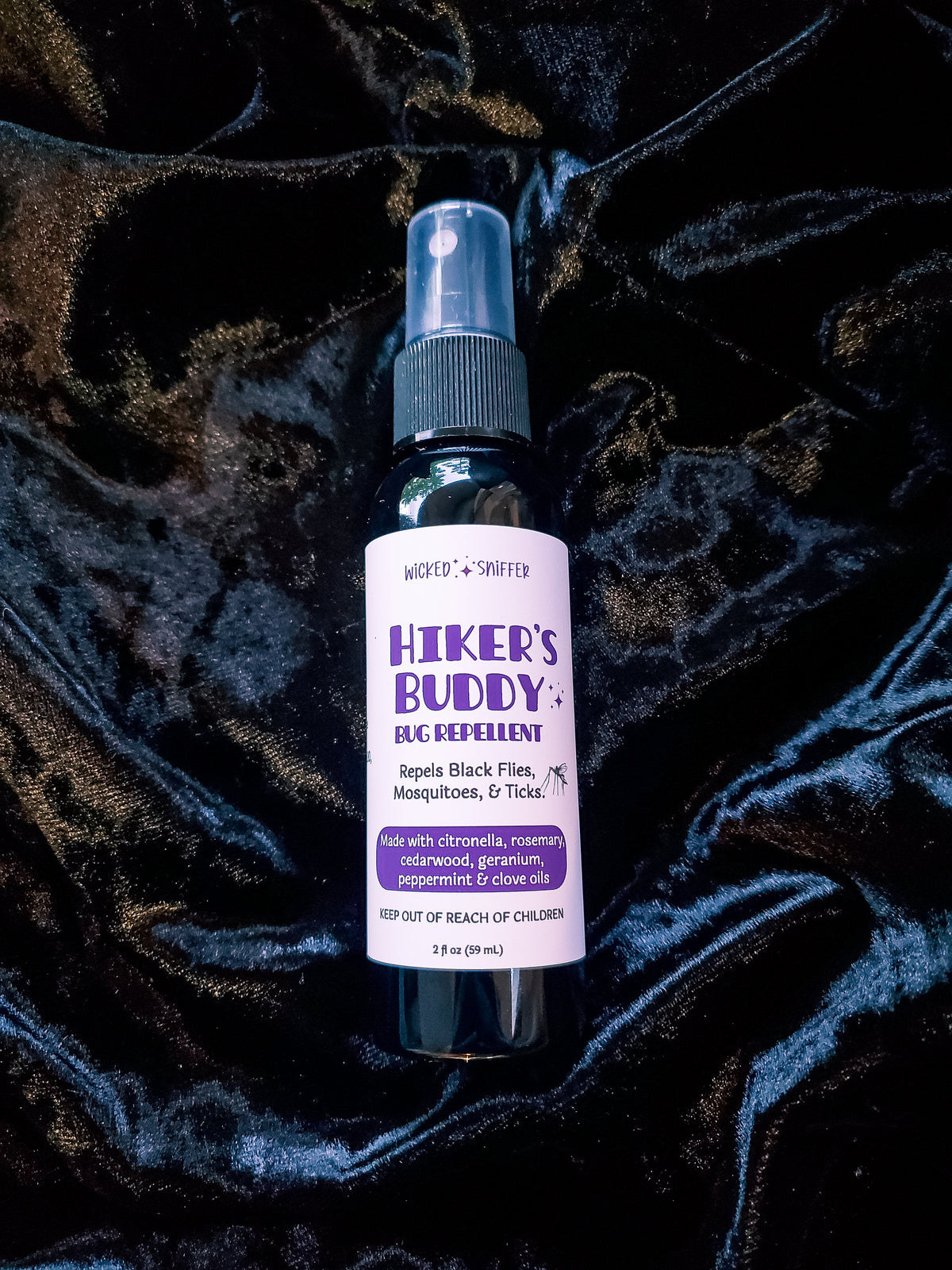 A bottle of Hiker's Buddy Bug Repellent with a white label and purple text, placed on a dark, textured velvet fabric background.