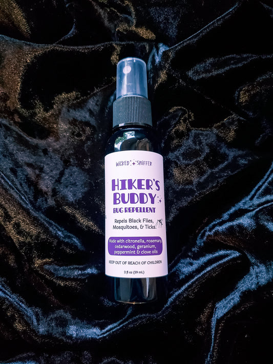 A bottle of Hiker's Buddy Bug Repellent with a white label and purple text, placed on a dark, textured velvet fabric background.