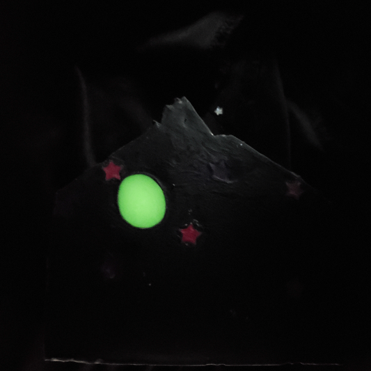 A black house-shaped soap with a glowing green oval and three pink star accents, shown against a very dark background.