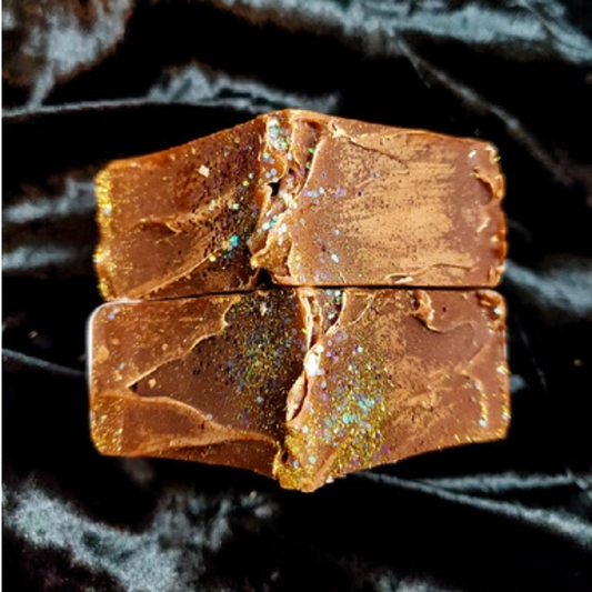 Two rectangular bars of brown soap with embedded gold glitter flakes, displayed on a black textured fabric background.