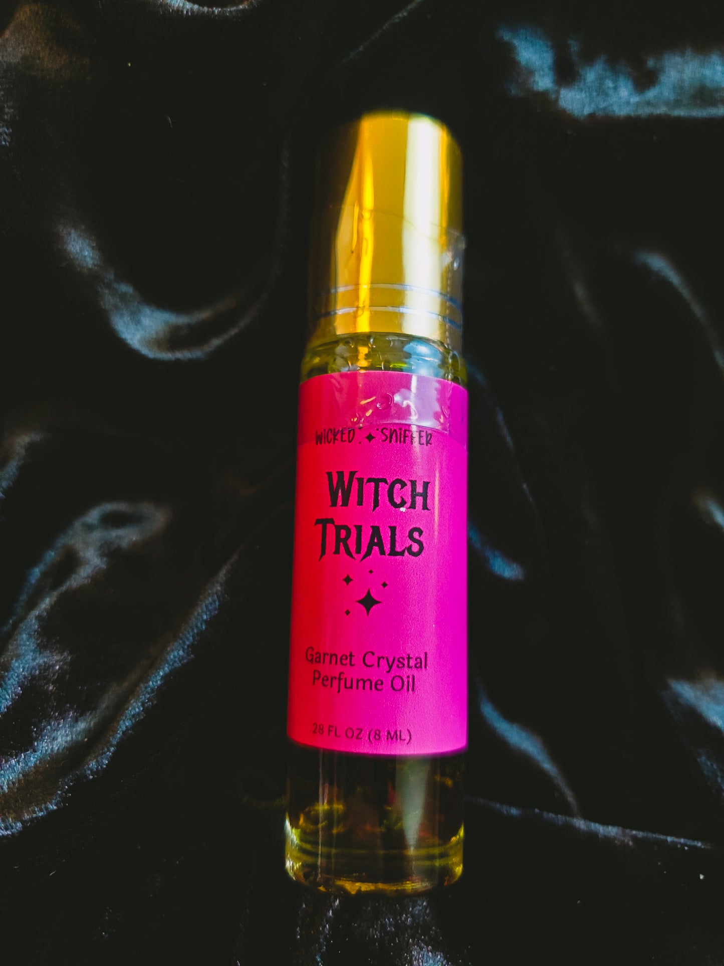 A bottle of 'Witch Trials' Garnet Crystal Perfume Oil with a bright pink label and gold cap, set against a dark, draped velvet background.